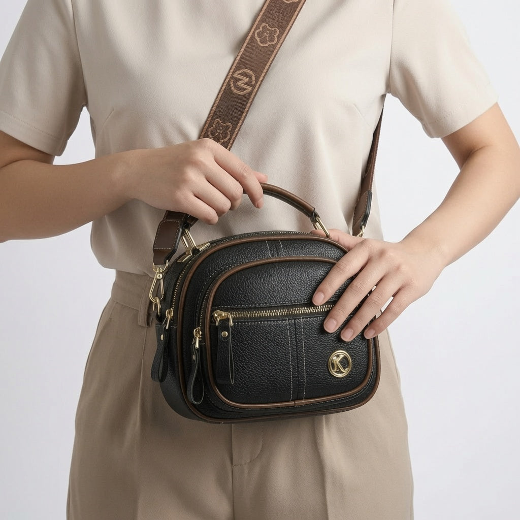 Bolso bandolera compacto de cuero negro con cremallera y correa estampada, placa K visible.