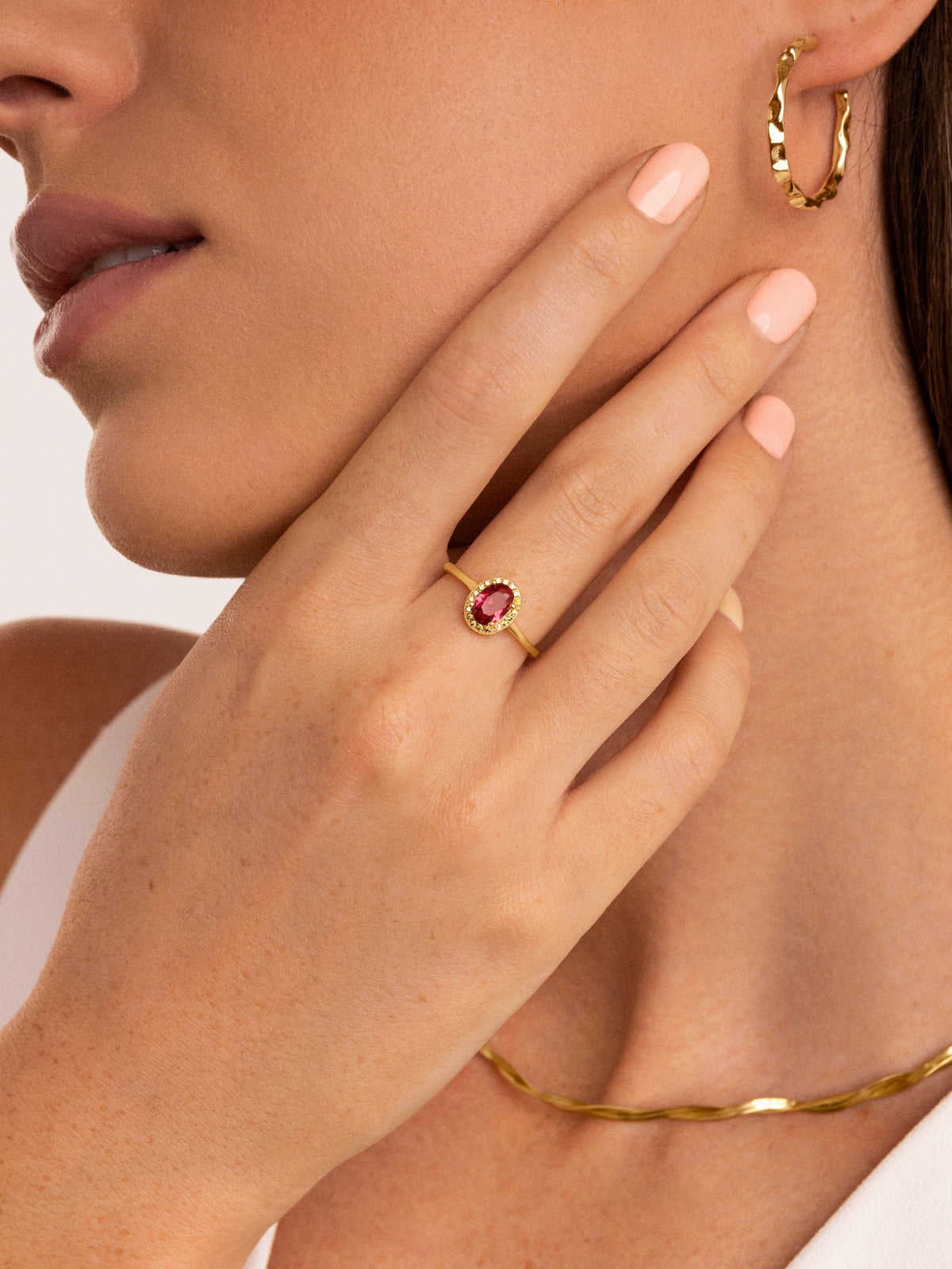 Anillo dorado con piedra roja ovalada y pedrería, sobre mano y uña